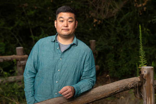 Taeyoon Lee, Ph.D. He is standing with his arm on a wooden rail. He is wearing a green button down shirt